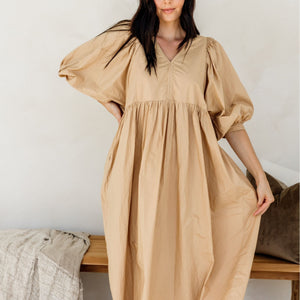 Woman wearing a beige dress sitting on a wooden bench against a white wall.