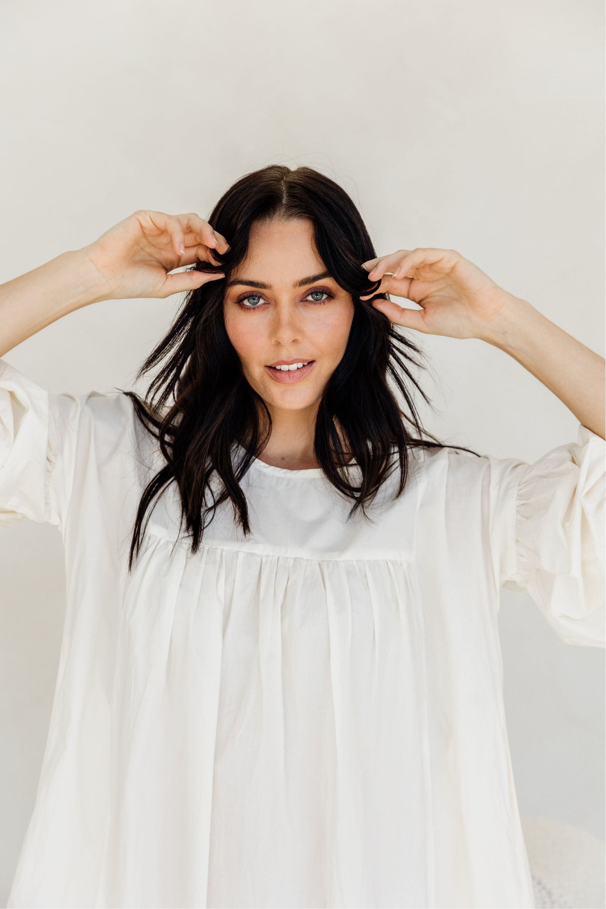 Woman in a white dress adjusting her hair against a plain background