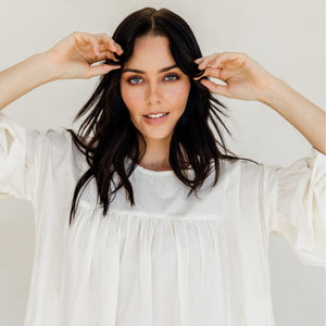 Woman in a white dress adjusting her hair against a plain background