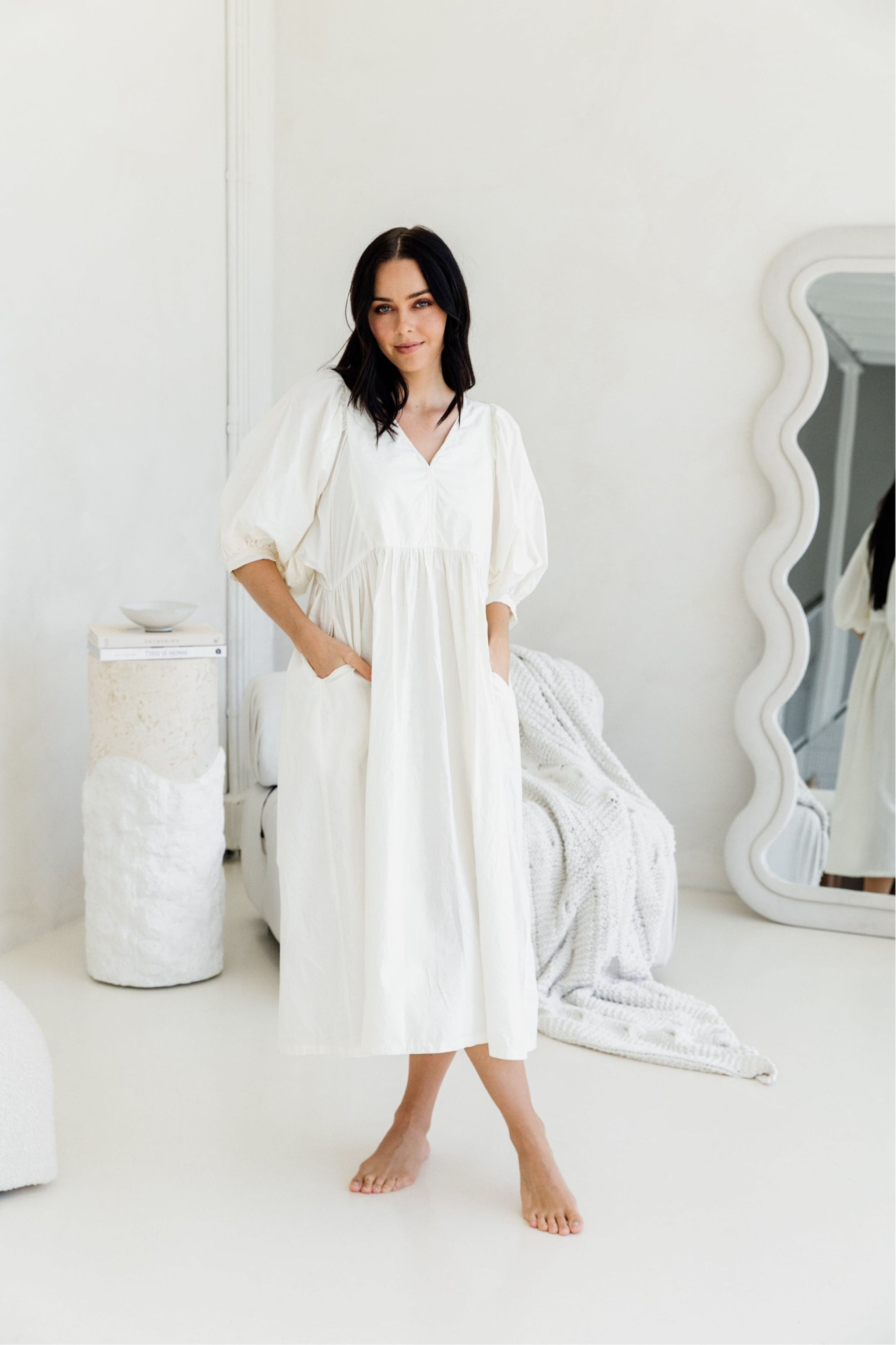 Woman wearing a white dress in a minimalistic room with a mirror and decorative items.