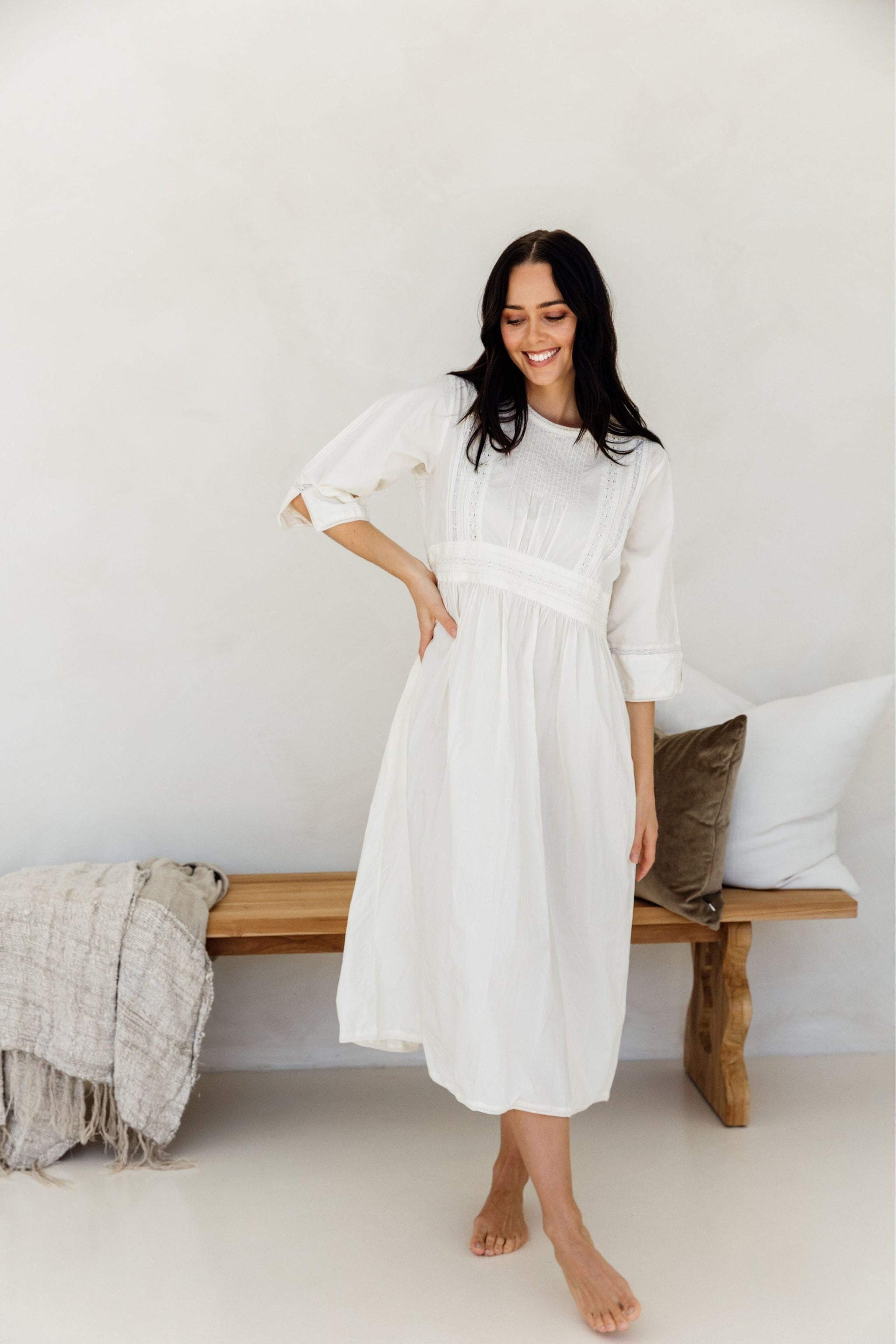 A person standing in a relaxed pose wearing a white mid-length dress with long sleeves and side pockets, in a room with a wooden bench and pillows in the background.