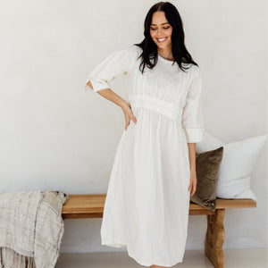 A person standing in a relaxed pose wearing a white mid-length dress with long sleeves and side pockets, in a room with a wooden bench and pillows in the background.