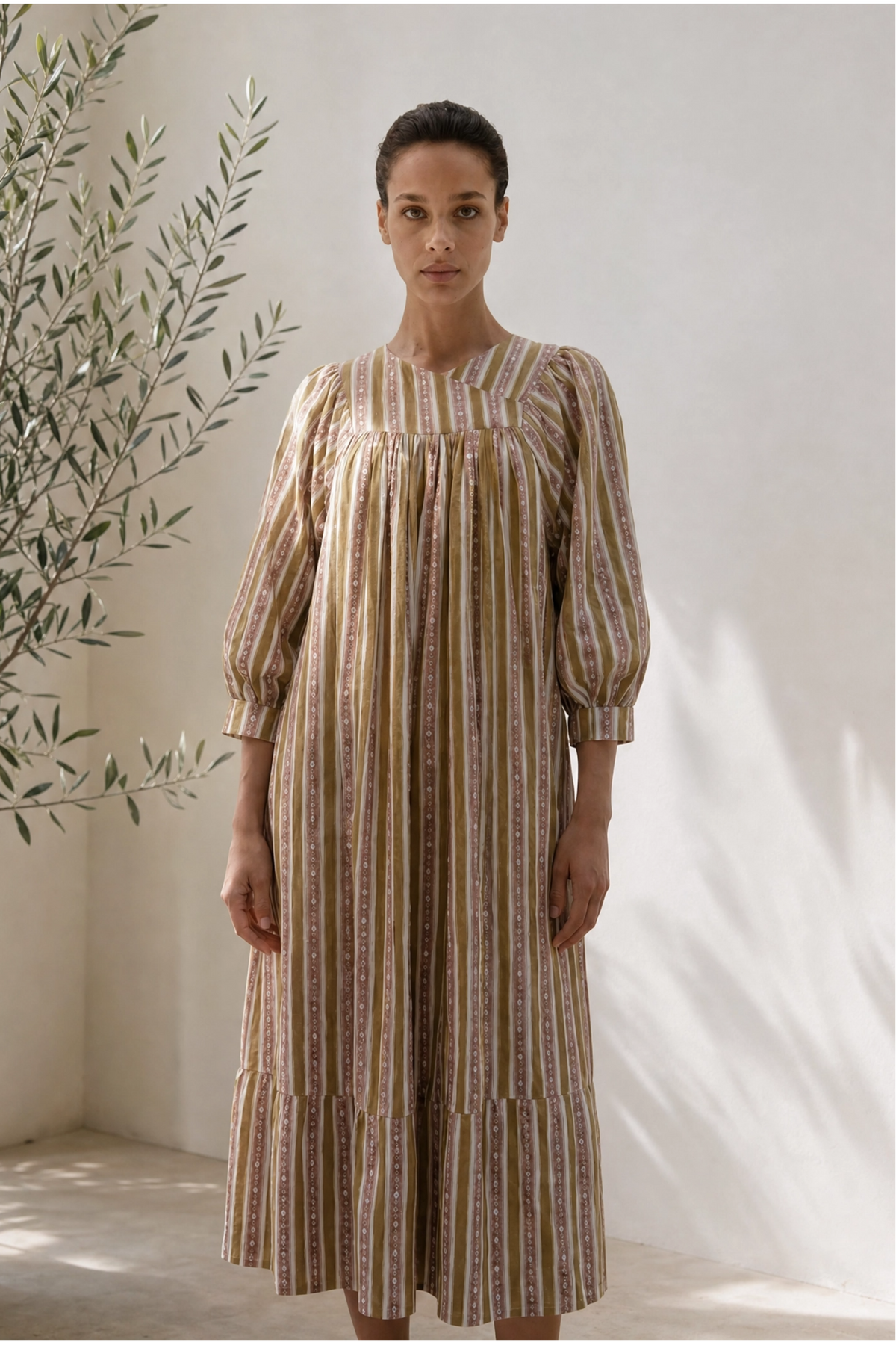 Woman wearing a striped dress standing against a white wall with a plant to the left.