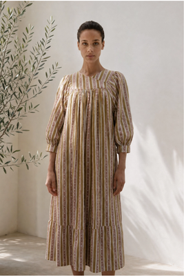 Woman wearing a striped dress standing against a white wall with a plant to the left.