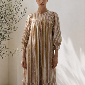 Woman wearing a striped dress standing against a white wall with a plant to the left.